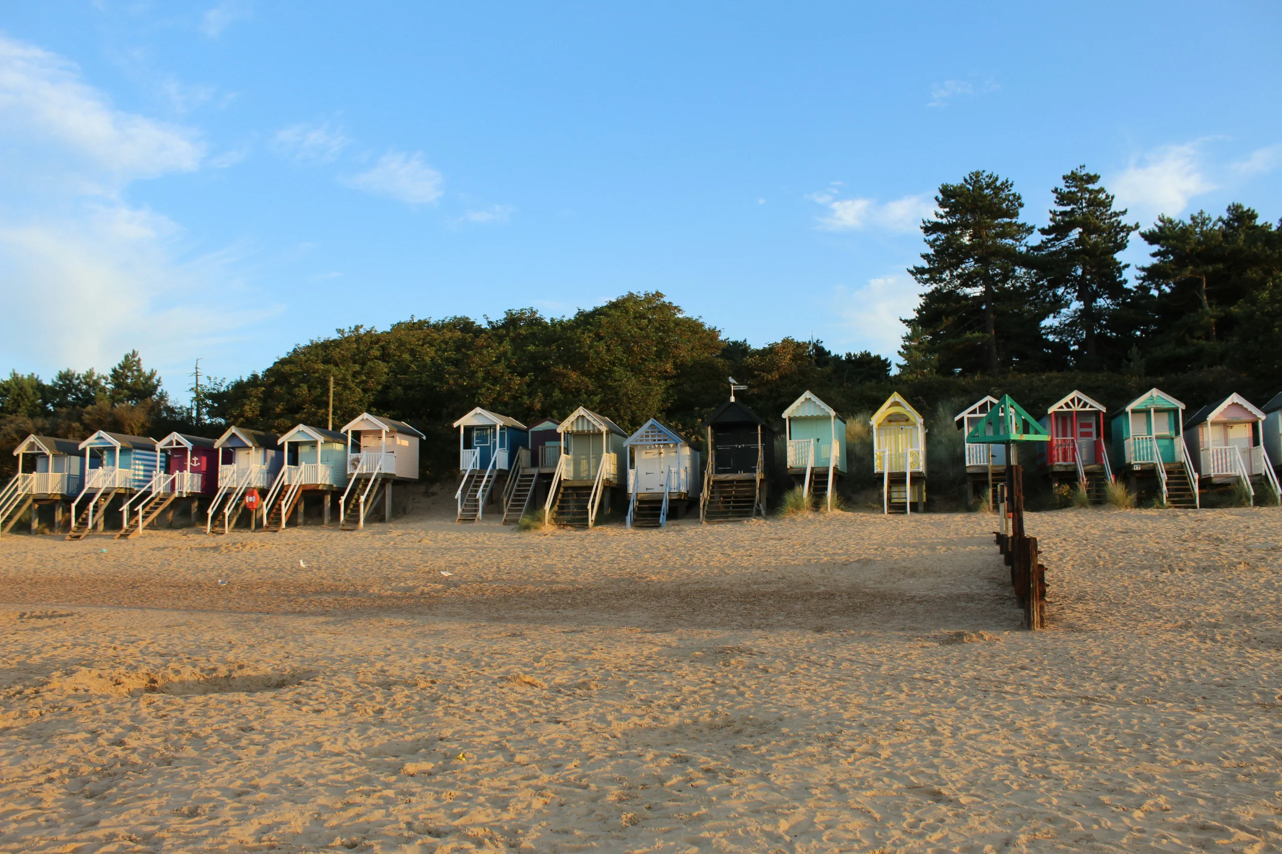 A row of colourful beach huts along the sand at Wells-next-the-Sea beach in North Norfolk, with pine woods behind
