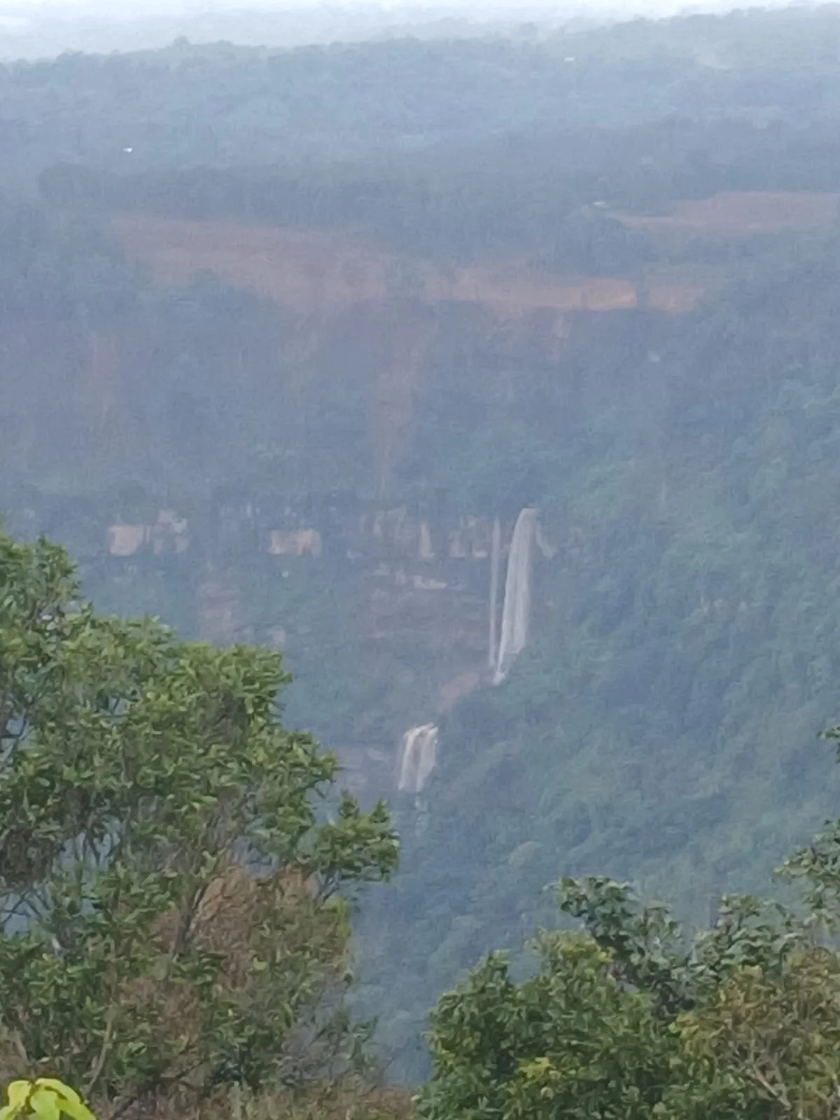 A distant waterfall falling down a forested cliff face, seen through haze from a viewpoint
