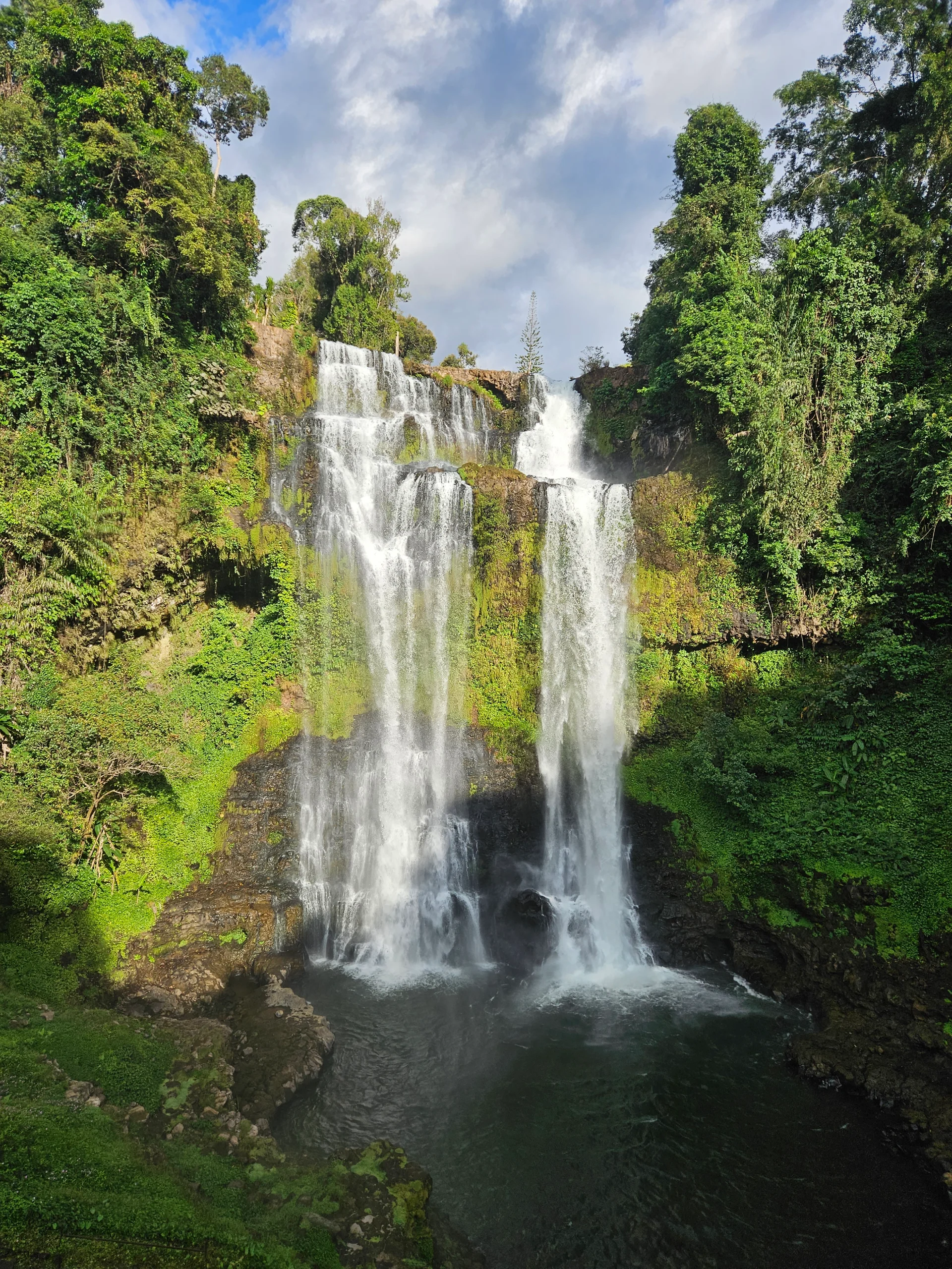 A wide multi-tiered waterfall spilling over mossy rock into a green plunge pool