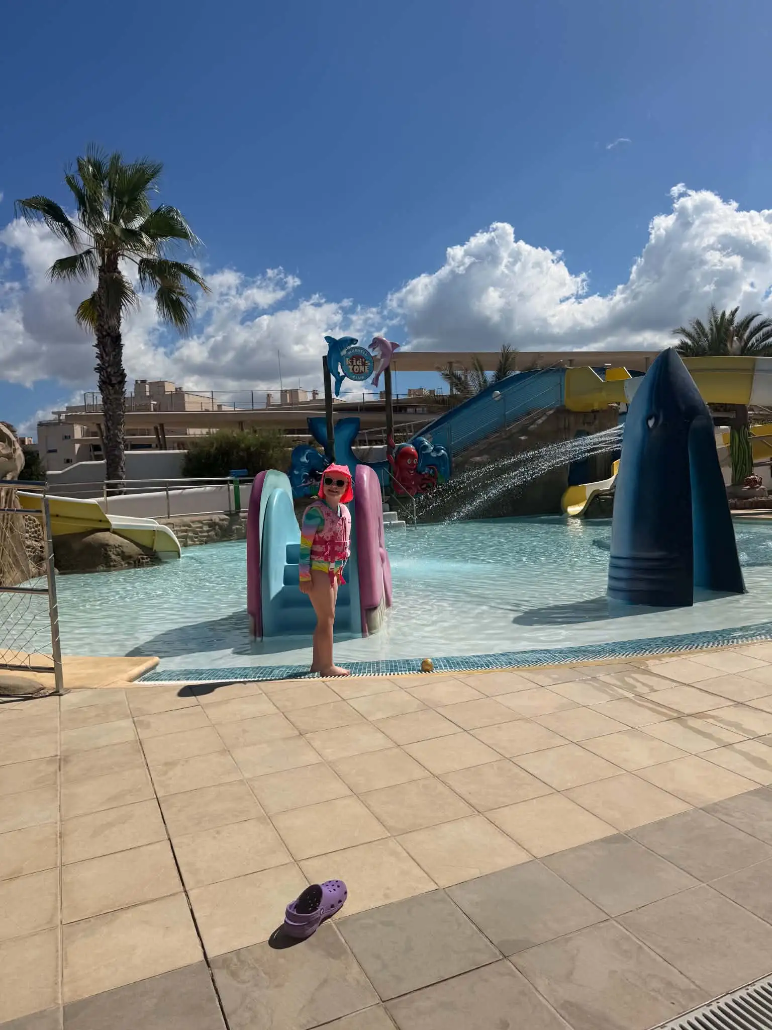 Young girl in floatation vest stood in front of hotel pool with water slides