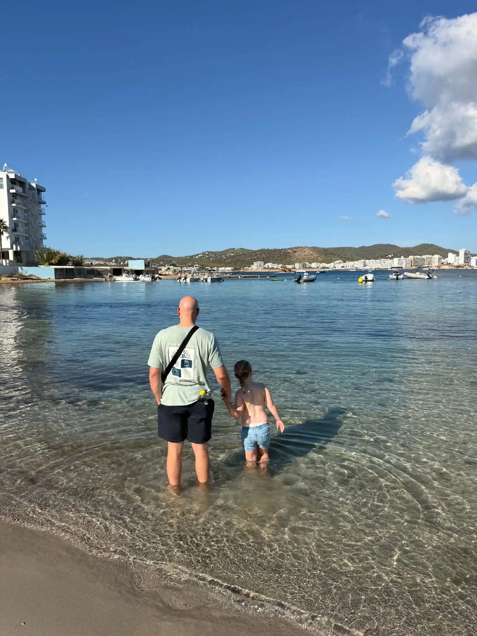 Dad and daughter holding hands in the sea facing away from the camera looking at some boats