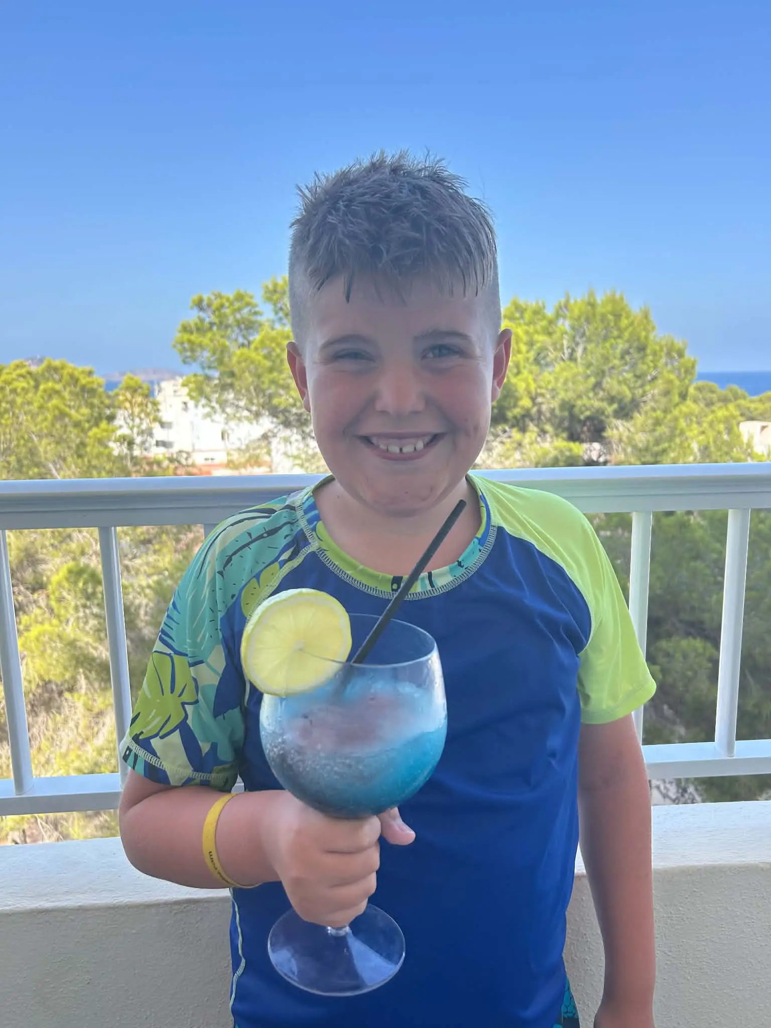A boy smiling on a balcony holding a large blue mocktail with a slice of lemon