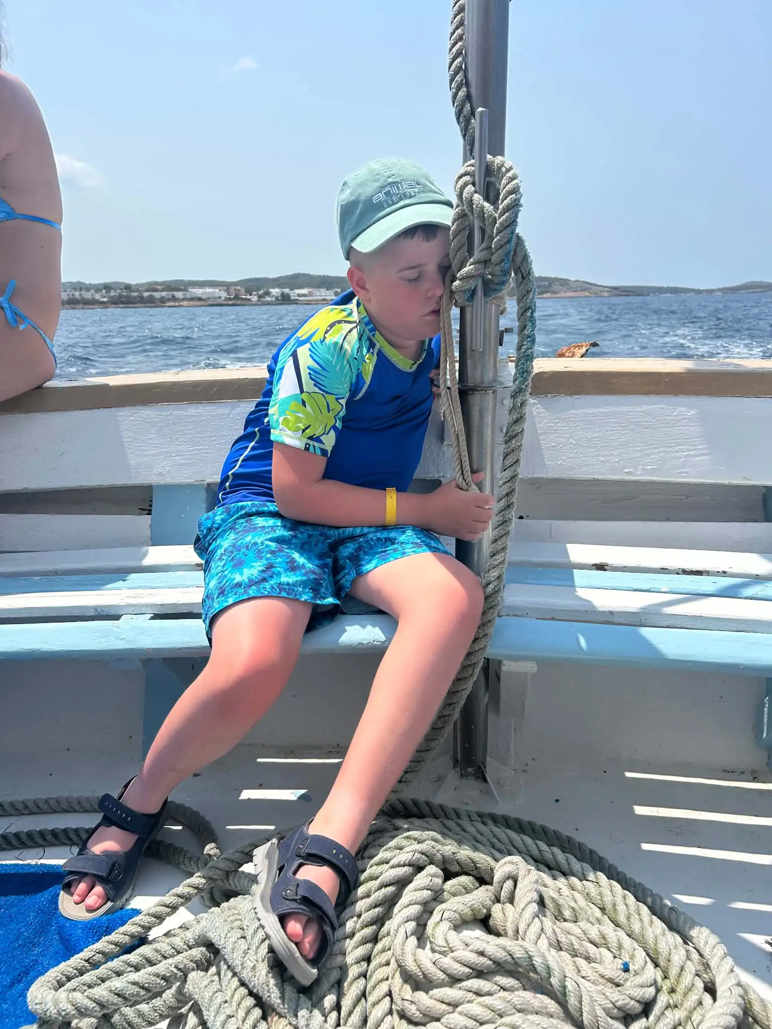 A boy in a blue rash vest and cap asleep against a post on the deck of a boat